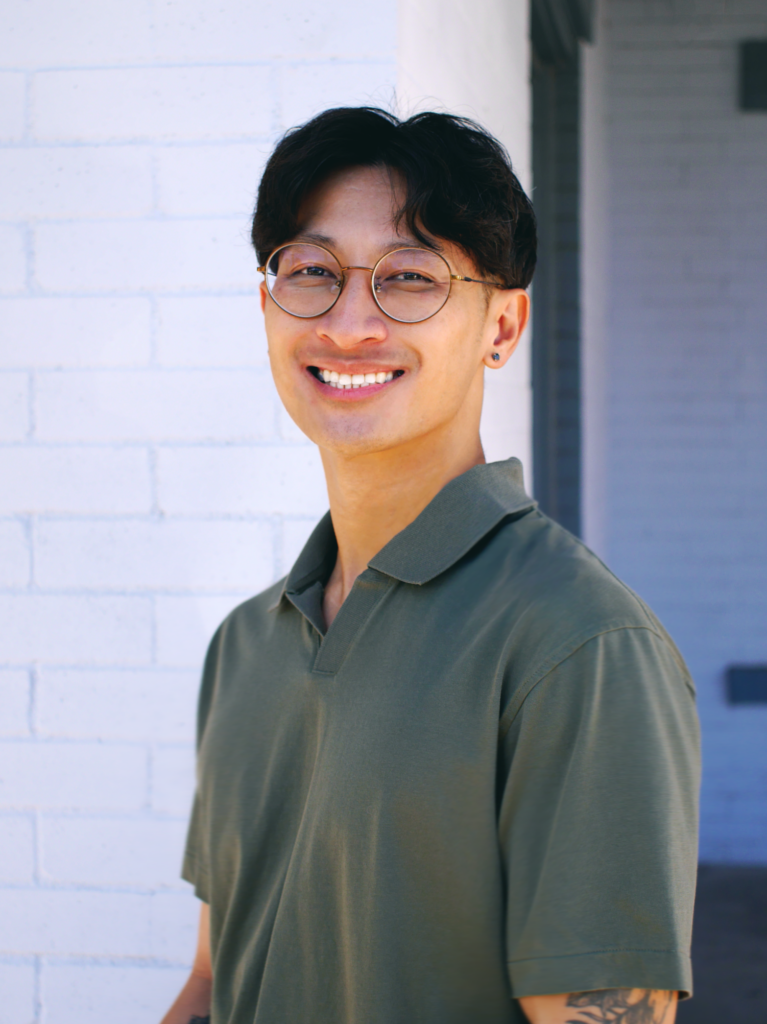 Headshot of a smiling man with short dark hair and glasses wearing an olive green collared shirt against a light brick wall background.